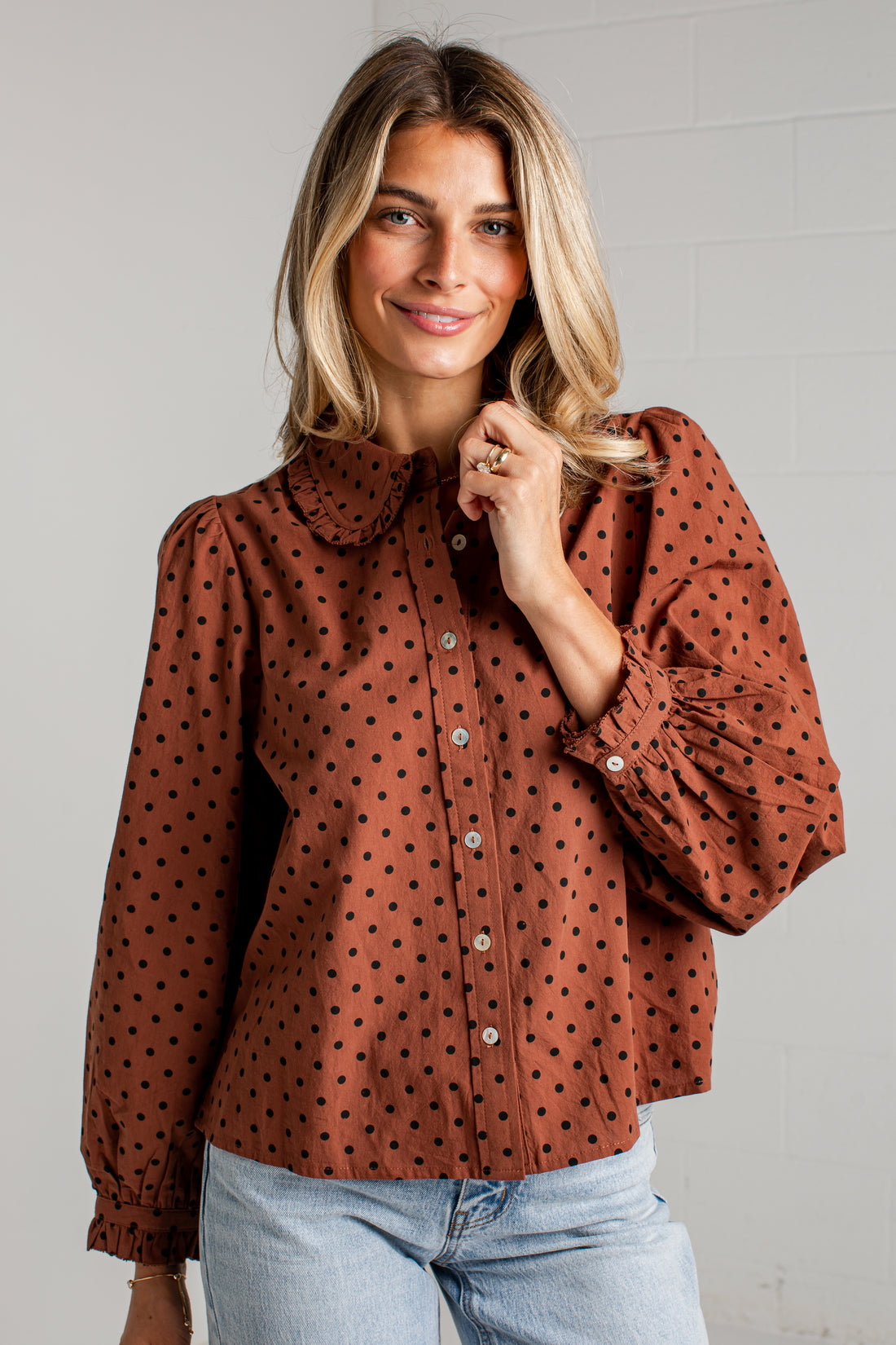 A woman wearing a rust-brown long-sleeve blouse with black polka dots, a Peter Pan collar, and voluminous ruffled sleeves, paired with light-wash blue jeans against a white brick wall.
