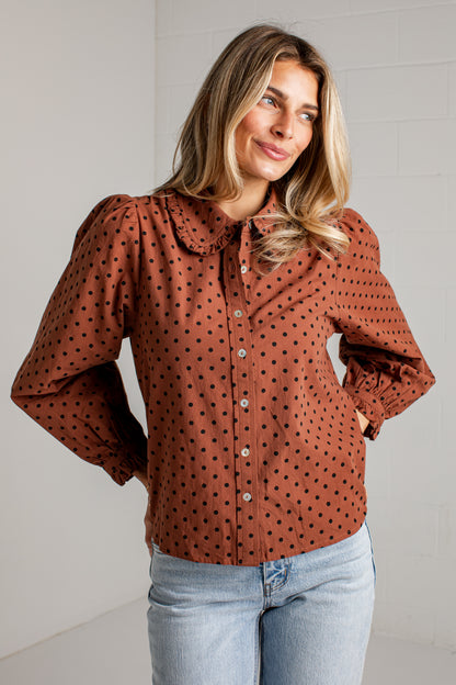 A woman wearing a rust-brown long-sleeve blouse with black polka dots and a large Peter Pan collar, paired with light-wash blue jeans, standing against a white brick wall.