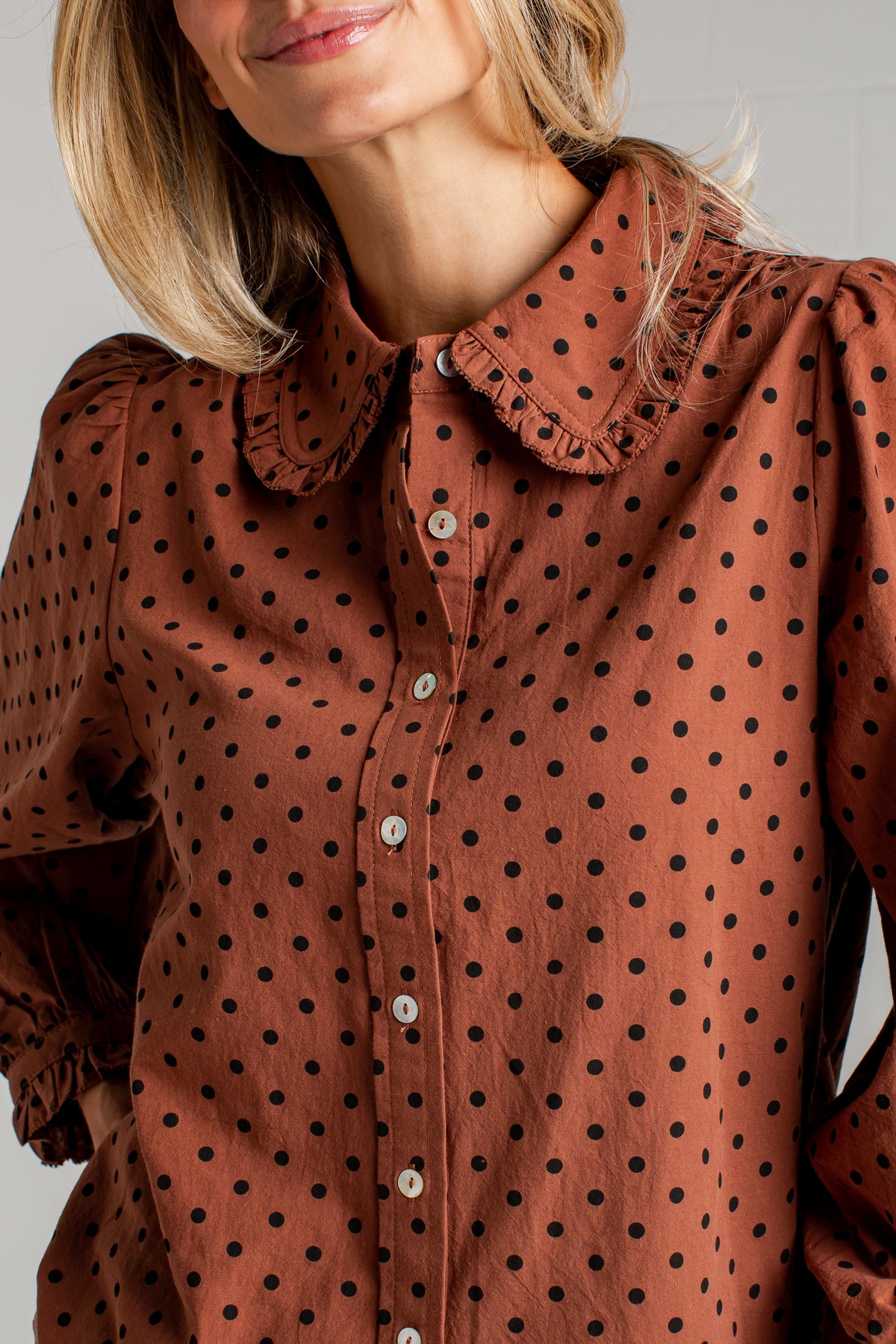 A close-up of a woman wearing a rust-brown long-sleeve blouse with black polka dots, featuring a ruffled Peter Pan collar and white buttons.