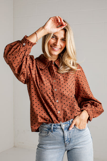 A smiling blonde woman wearing a rust-brown long-sleeve button-up blouse with black polka dots and light-wash blue jeans, posing in a bright indoor setting.