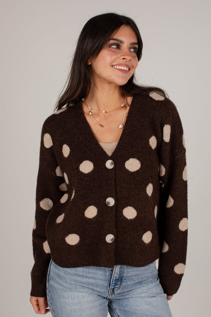 A woman wearing a dark brown knit cardigan with beige polka dots and silver buttons, paired with light blue jeans, against a plain light gray background.