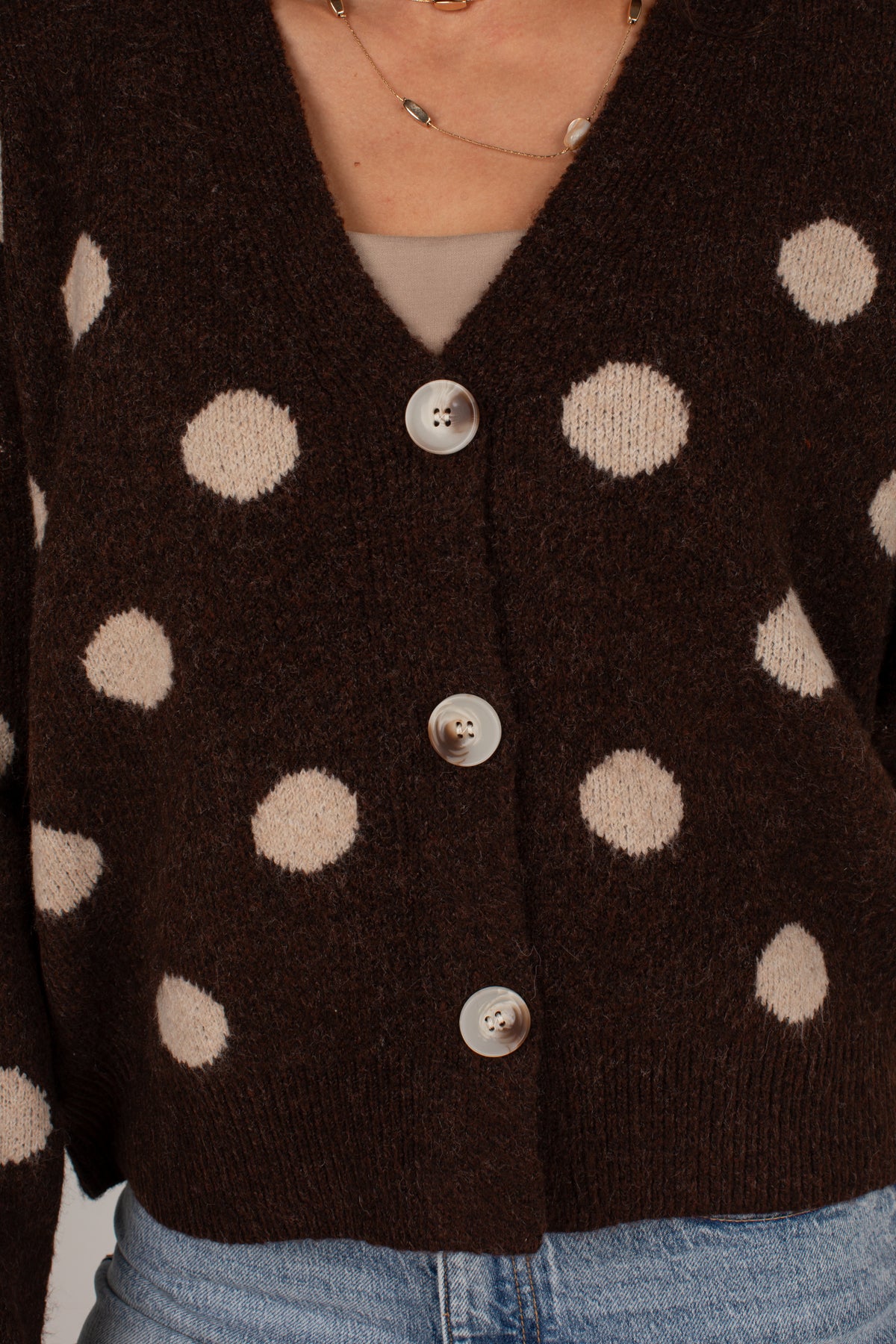 A close-up of a dark brown knit cardigan with large beige polka dots and silver buttons, worn by a model over a beige top and blue jeans.
