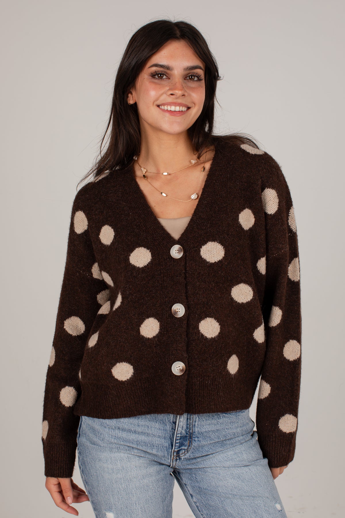 A woman wearing a dark brown fuzzy cardigan with large beige polka dots and light blue jeans, standing against a plain light gray studio background.