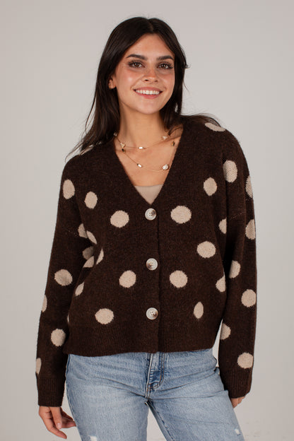 A woman wearing a dark brown fuzzy cardigan with large beige polka dots and light blue jeans, standing against a plain light gray studio background.