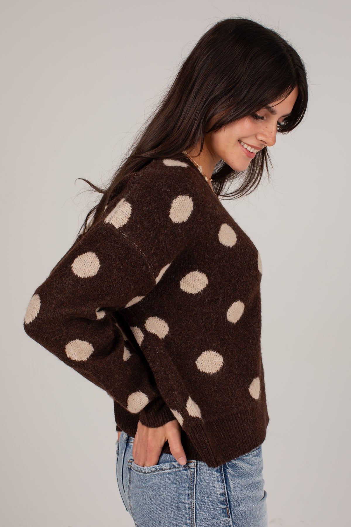 A woman wearing a brown fuzzy knit sweater with large beige polka dots, paired with light blue jeans, photographed against a plain light gray background.