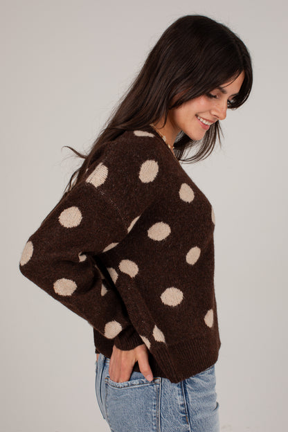 A woman wearing a brown fuzzy knit sweater with large beige polka dots, paired with light blue jeans, photographed against a plain light gray background.
