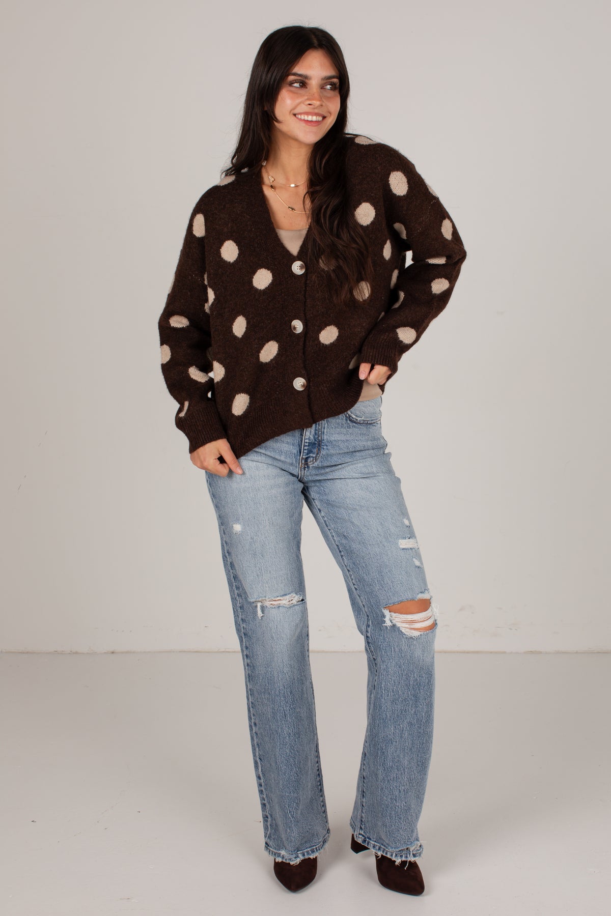 A woman modeling a dark brown button-up cardigan with large beige polka dots, paired with light-wash distressed jeans and dark brown pointed shoes, against a plain light gray studio background.