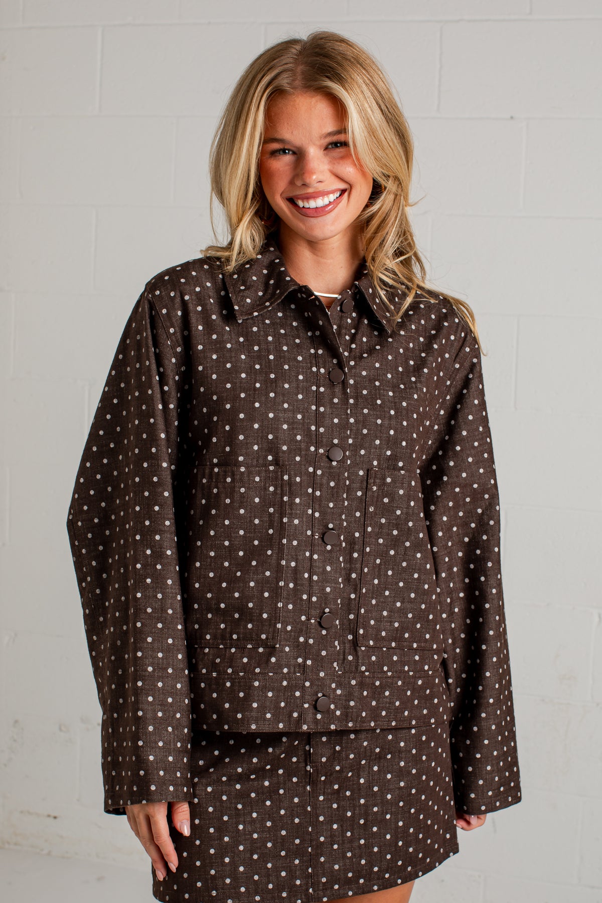 A smiling blonde woman wearing a brown long-sleeve button-up shirt and matching shorts set, both featuring a small white polka dot pattern, posed against a white painted brick wall.