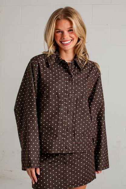 A smiling blonde woman wearing a brown long-sleeve button-up shirt and matching shorts set, both featuring a small white polka dot pattern, posed against a white painted brick wall.