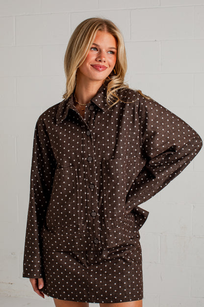 A woman modeling a dark brown long-sleeve button-up shirt dress with a white polka dot pattern, standing against a white brick wall.