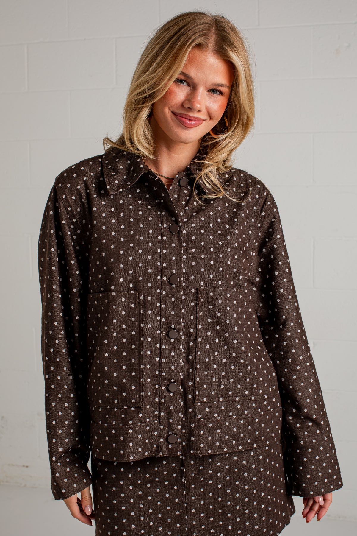 A woman wearing a dark brown long-sleeve button-up shirt with a white polka dot pattern, posed against a light gray brick wall.