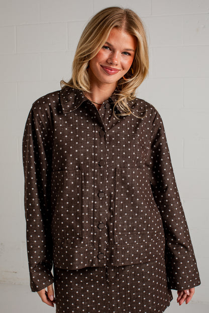 A woman wearing a dark brown long-sleeve button-up shirt with a white polka dot pattern, posed against a light gray brick wall.
