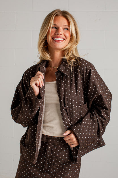 A smiling blonde woman wearing a brown and white polka dot jacket over a beige tank top, standing against a white brick wall.