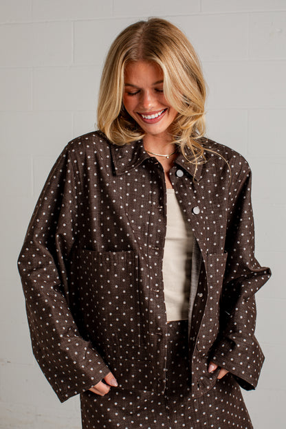 A woman wearing a brown button-up shirt jacket with a white polka dot pattern, layered over a cream top, standing against a white brick wall.