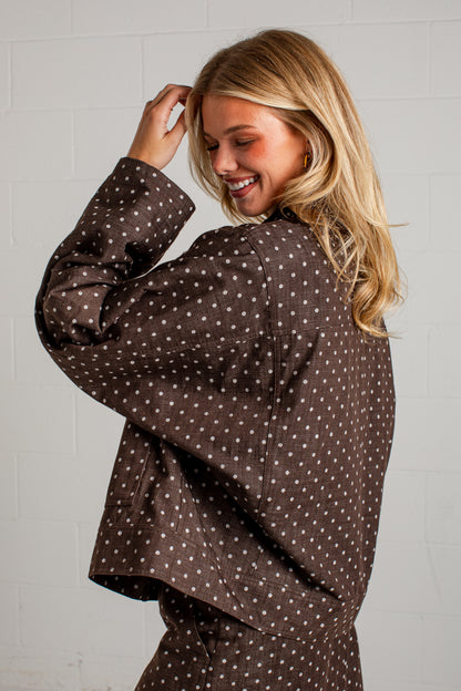 A woman with blonde hair is shown from the back wearing a dark brown long-sleeve top with a small white polka dot pattern, posing against a light gray brick wall.