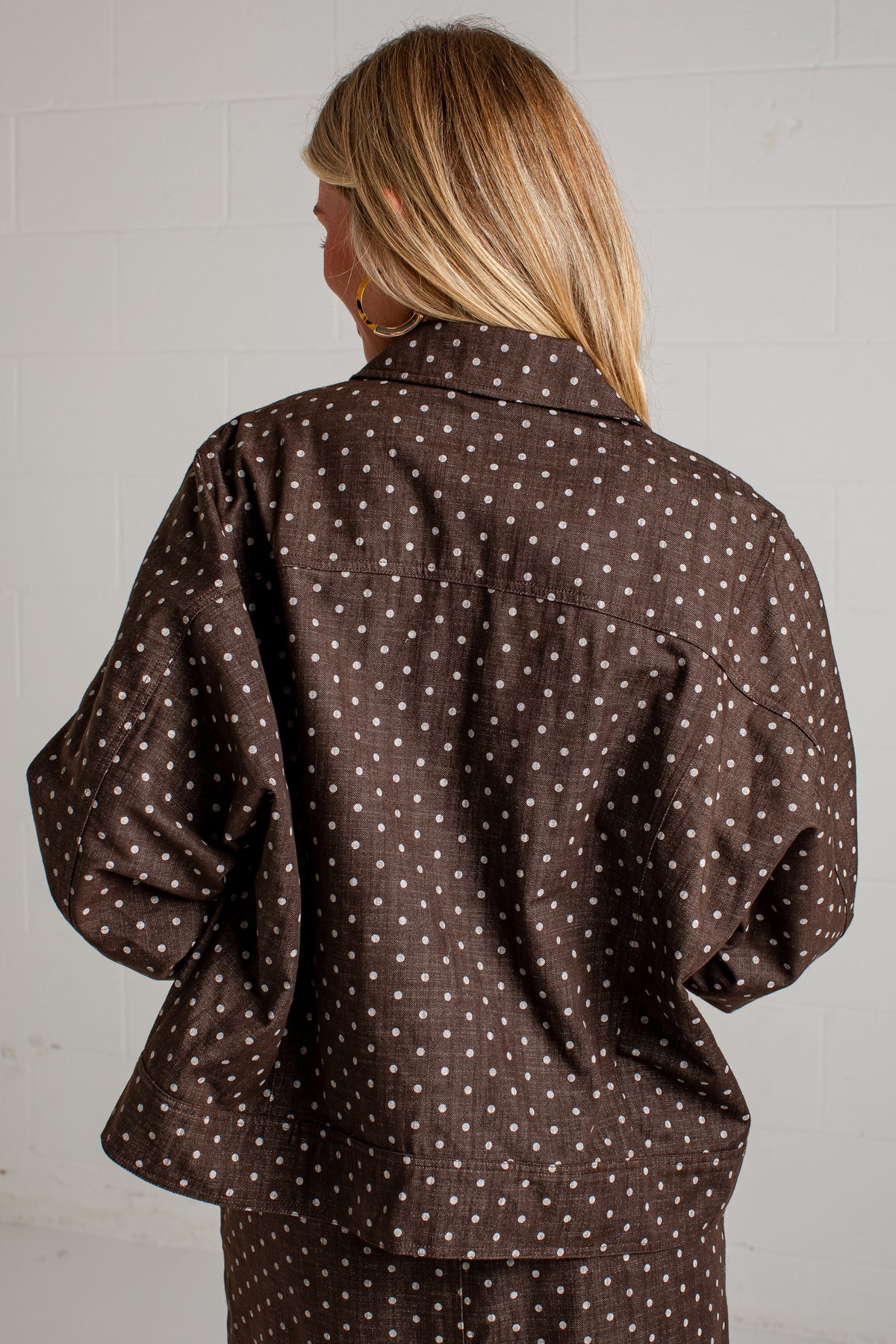 A woman viewed from behind wearing a dark brown button-up shirt with a white polka dot pattern, set against a light gray brick wall.