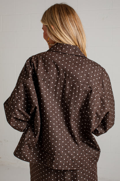 A woman viewed from behind wearing a dark brown button-up shirt with a white polka dot pattern, set against a light gray brick wall.