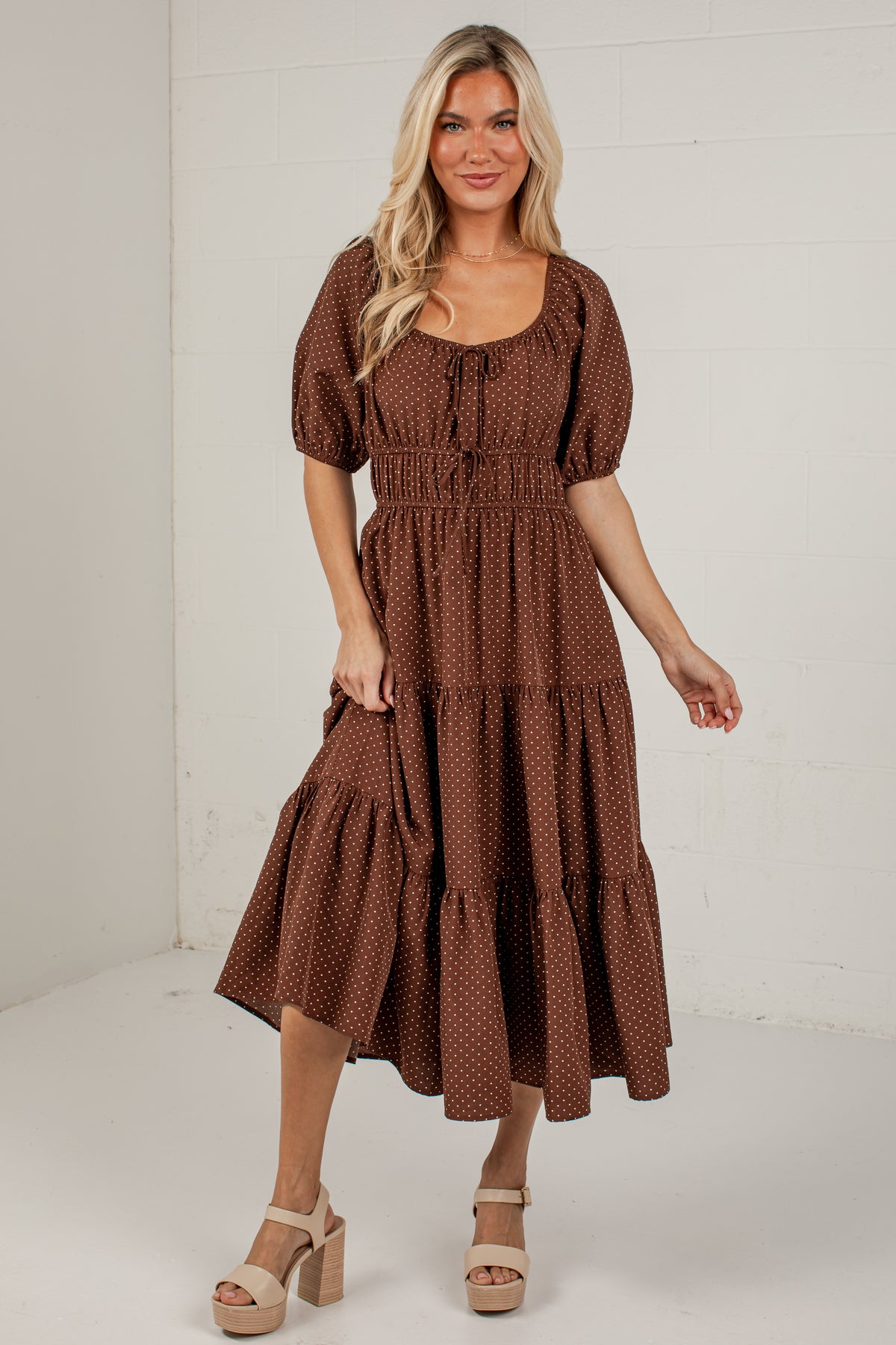 A woman modeling a brown tiered midi dress with a small white polka dot pattern, puff sleeves, and a tie neckline, standing against a white paneled wall.