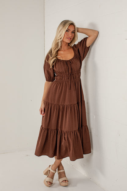 A woman modeling a brown tiered midi dress with a small white polka dot pattern, standing against a white wall.