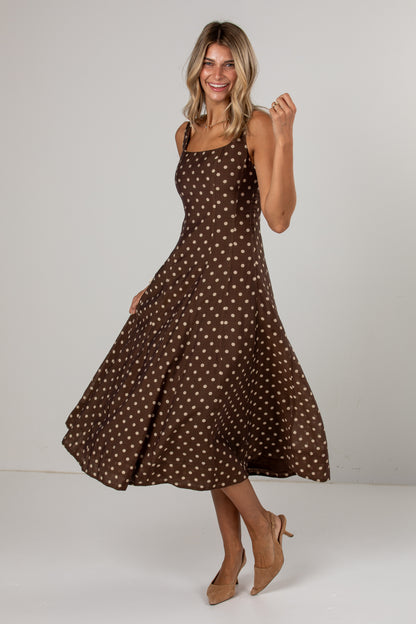 A woman modeling a brown midi dress with a beige polka dot pattern, standing in a studio setting against a light gray background.