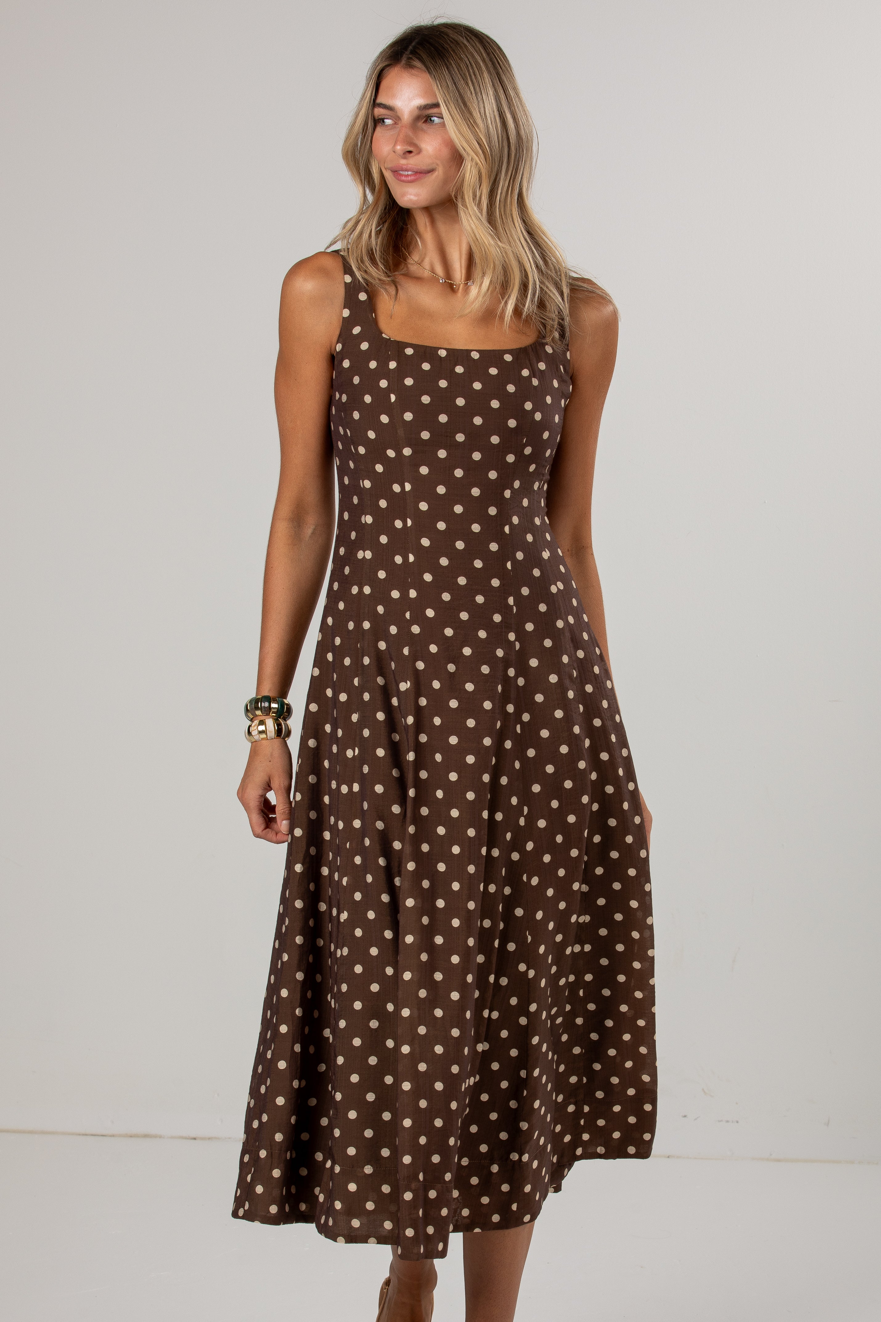 A woman modeling a brown sleeveless midi dress with a white polka dot pattern against a plain light gray studio background.