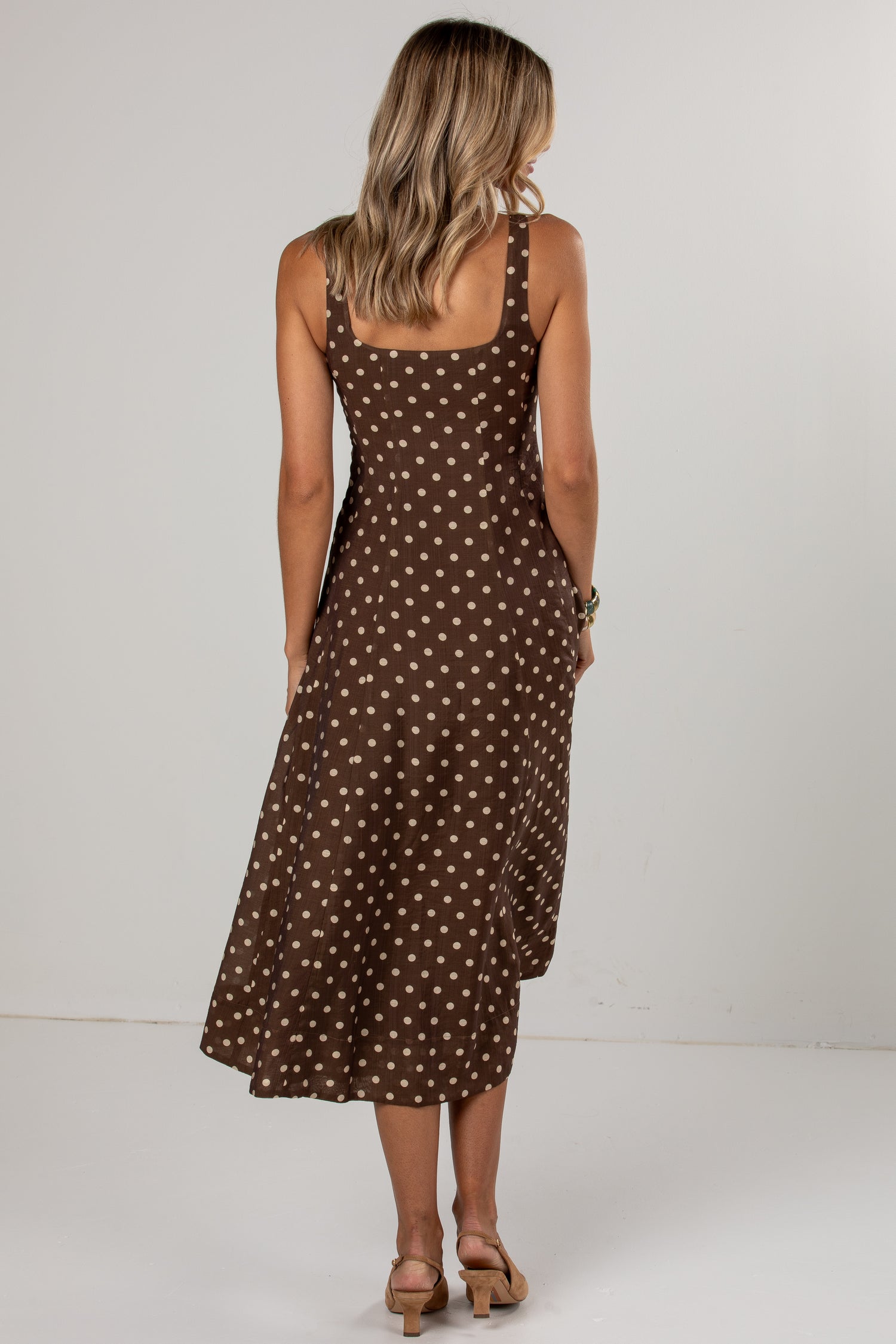 A woman viewed from behind wearing a brown midi dress with a white polka dot pattern, paired with tan heeled sandals, set against a plain light gray studio background.