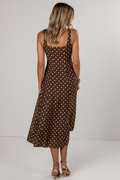 A woman viewed from behind wearing a brown midi dress with a white polka dot pattern, paired with tan heeled sandals, set against a plain light gray studio background.