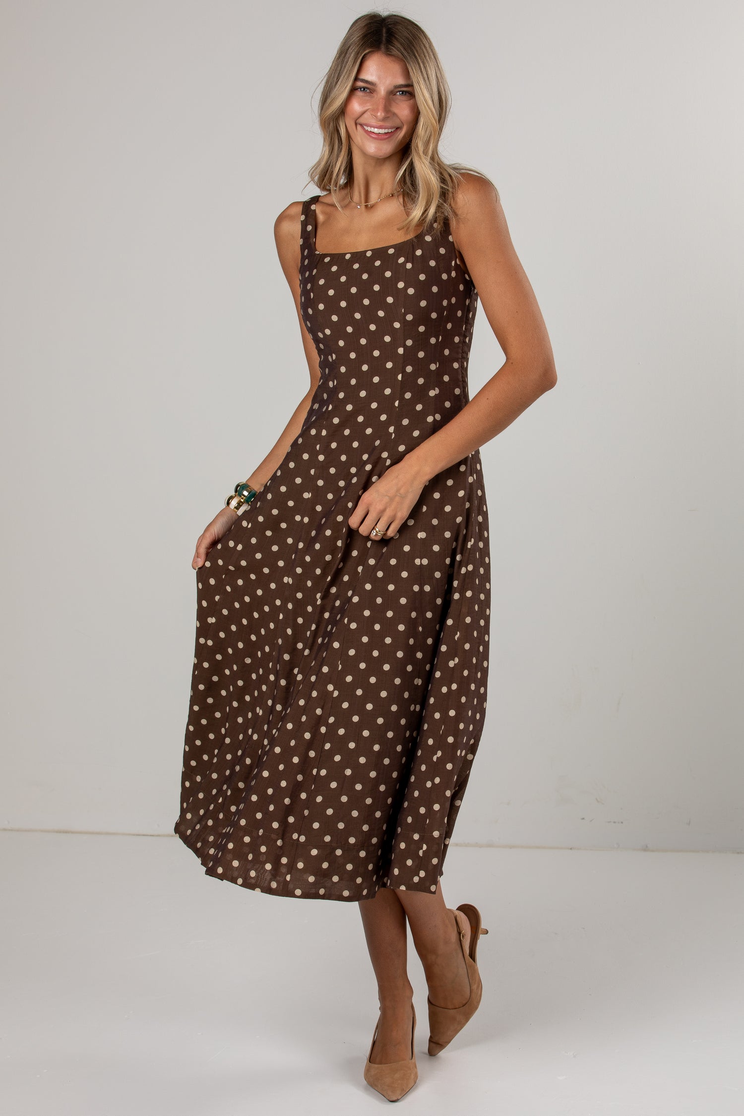 A woman modeling a brown midi dress with a white polka dot pattern, photographed against a neutral light gray studio background.