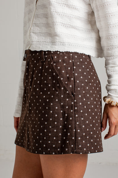A close-up of a person wearing a brown mini skirt with a white polka dot pattern, paired with a white textured knit top.