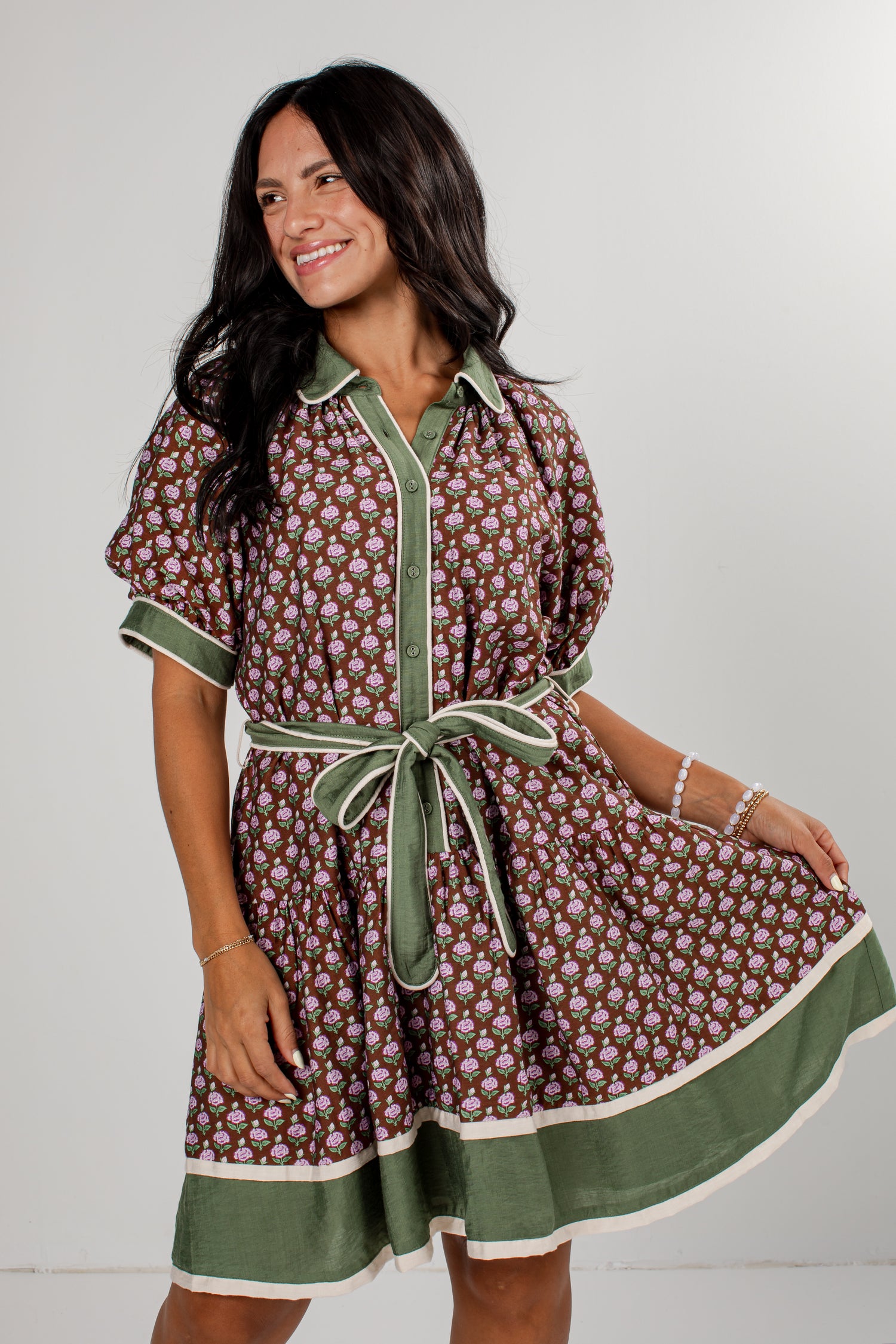 A woman modeling a short-sleeve button-front dress with a brown and beige floral print, olive green trim, and a matching green tie belt, set against a plain light gray studio background.