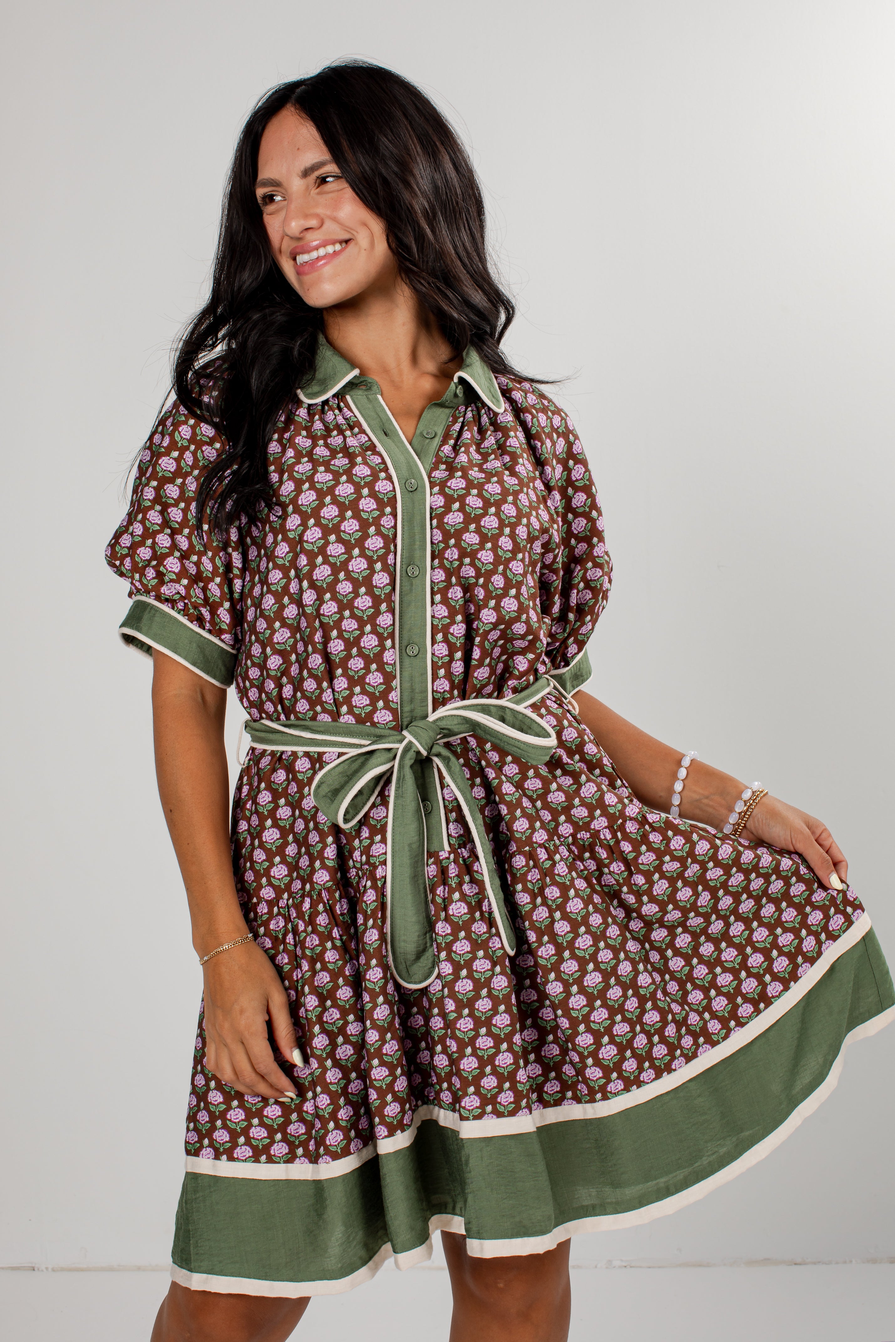 A woman modeling a short-sleeve button-front dress with a brown and beige floral print, olive green trim, and a matching green tie belt, set against a plain light gray studio background.