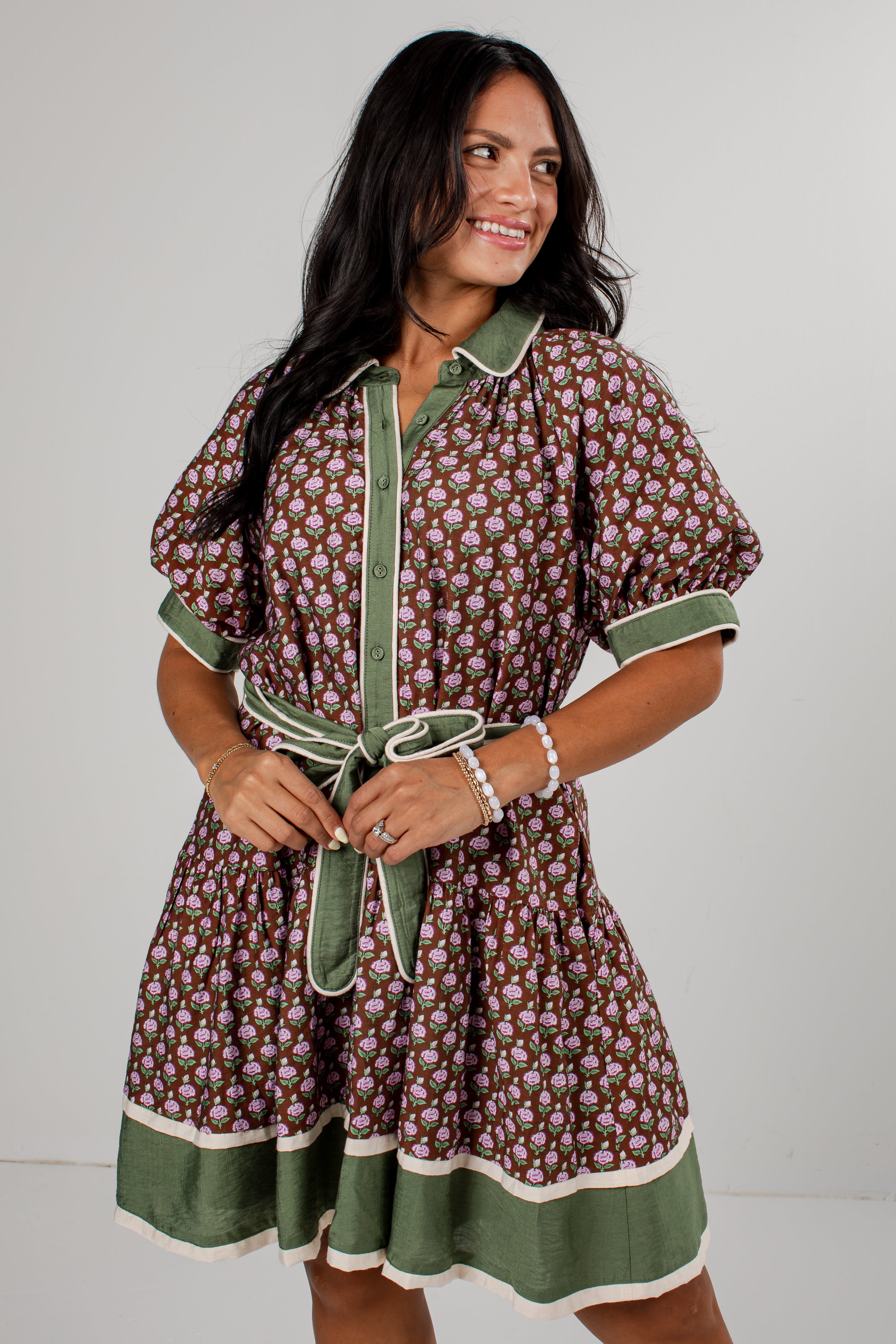 A woman modeling a short-sleeve button-front dress with a brown and beige floral print, olive green trim, and a matching green tie belt, set against a plain light gray studio background.
