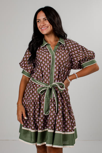 A woman modeling a short-sleeve button-front dress with a brown and beige floral print, olive green trim, and a matching green tie belt, set against a plain light gray studio background.