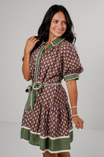 A woman modeling a short-sleeve button-front dress with a brown and beige floral print, olive green trim, and a matching green tie belt, set against a plain light gray studio background.