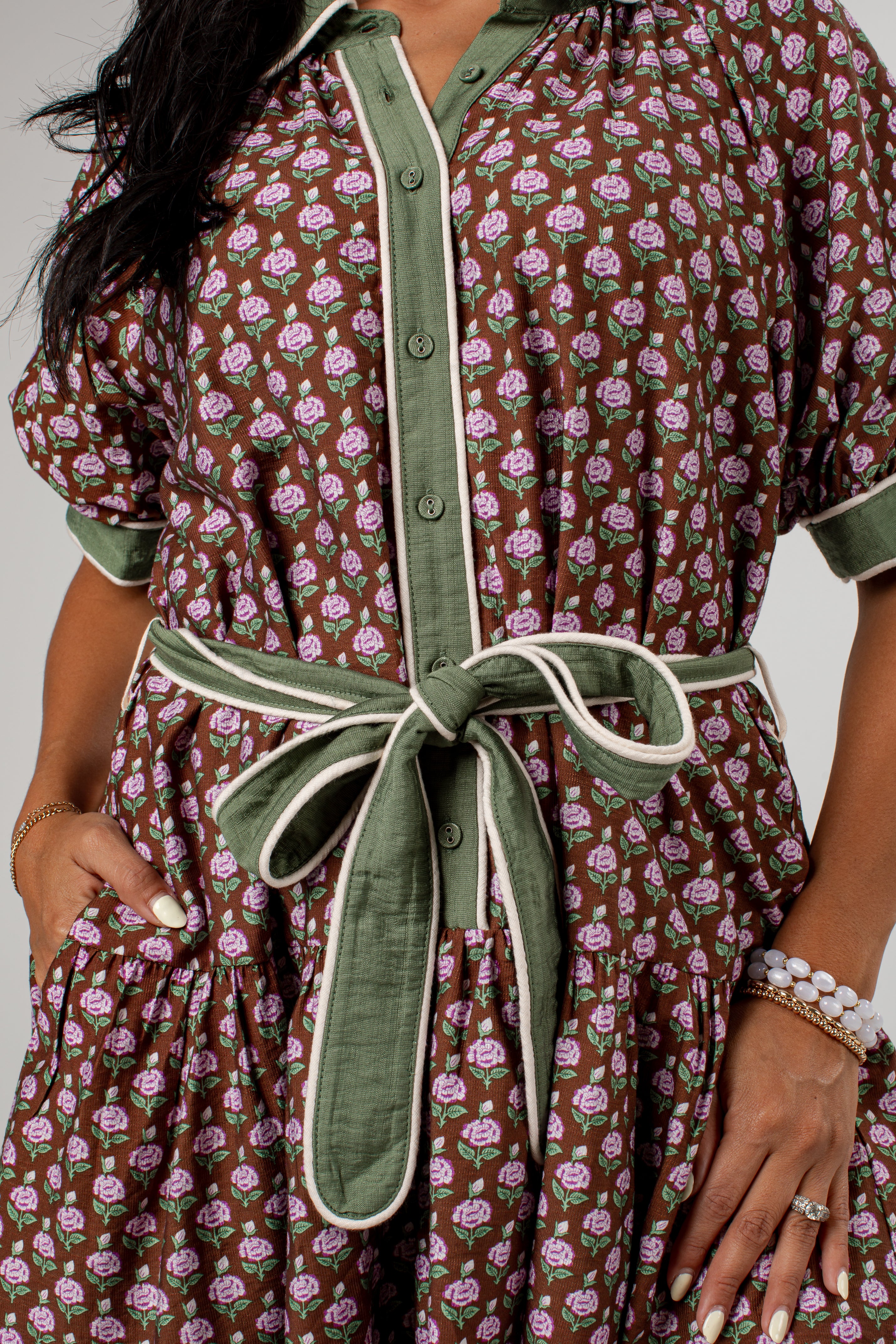 A close-up of a woman wearing a brown short-sleeve shirt dress with a pink and green floral print, featuring olive green trim and a matching green tie belt.