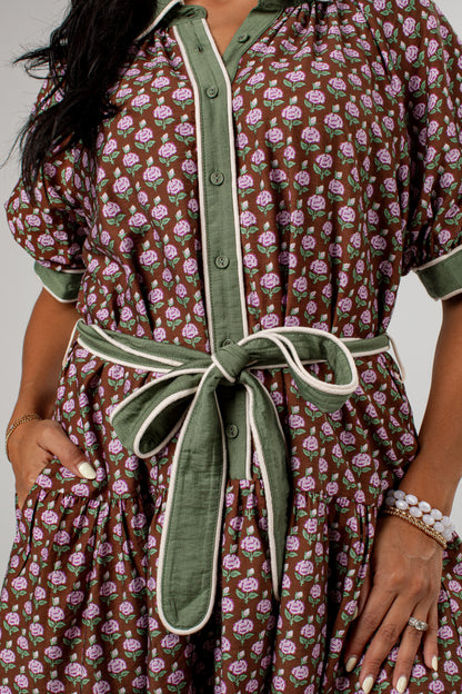 A close-up of a woman wearing a brown short-sleeve shirt dress with a pink and green floral print, featuring olive green trim and a matching green tie belt.