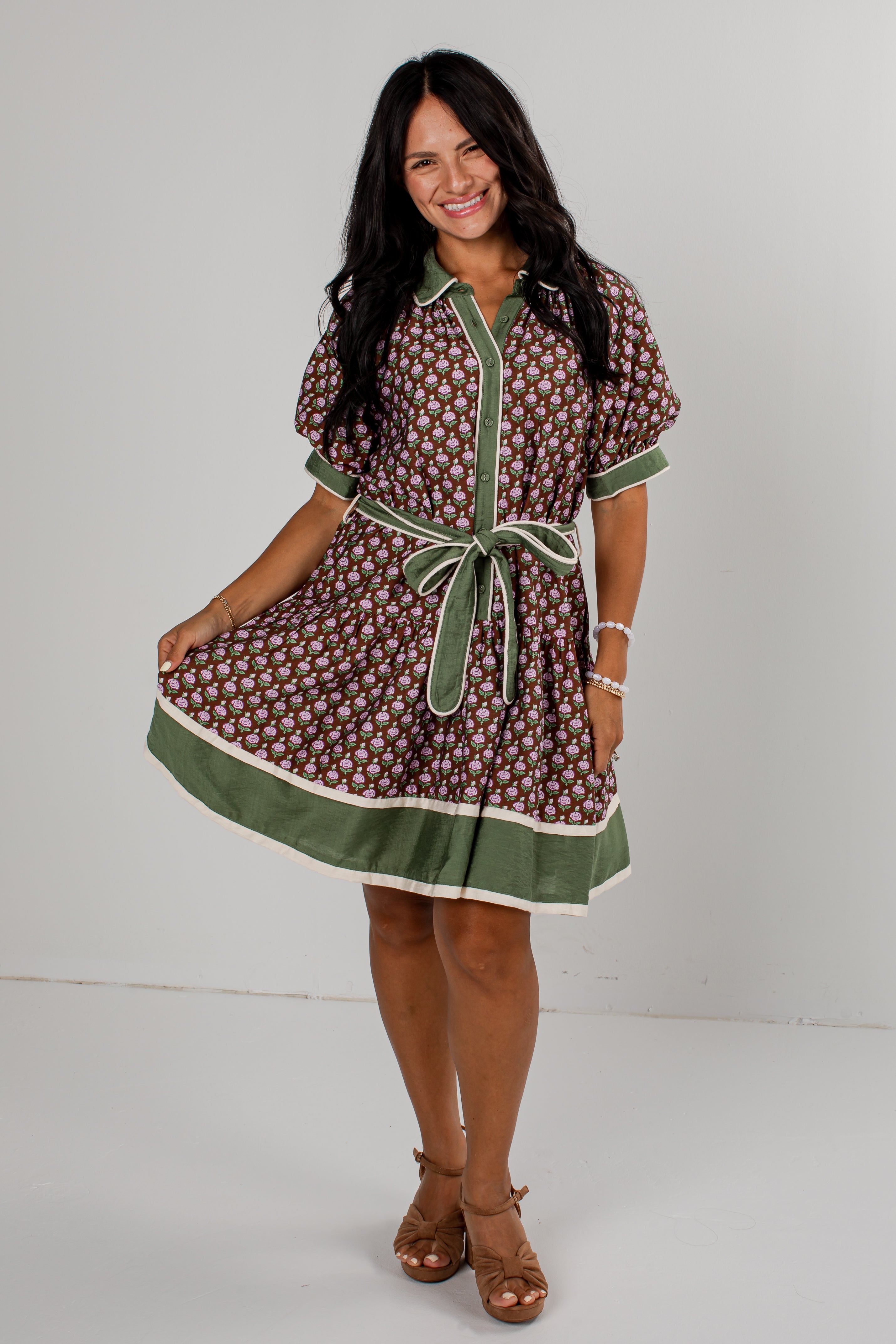 A woman modeling a short-sleeve button-front dress with an allover brown and white floral pattern, olive green trim, and a matching green tie belt, photographed against a plain light gray studio background.
