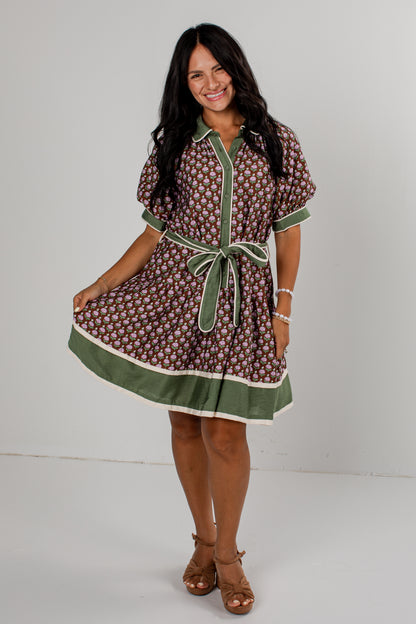 A woman modeling a short-sleeve button-front dress with an allover brown and white floral pattern, olive green trim, and a matching green tie belt, photographed against a plain light gray studio background.