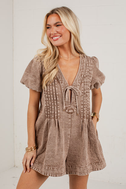A woman modeling a short-sleeve romper in a textured taupe-brown fabric with a tie-front detail and vertical stitched accents, posed against a white brick wall.