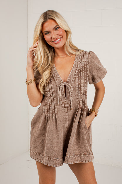 A woman modeling a short-sleeve brown romper with a tie-front neckline and vertical textured detailing, standing against a white brick wall.