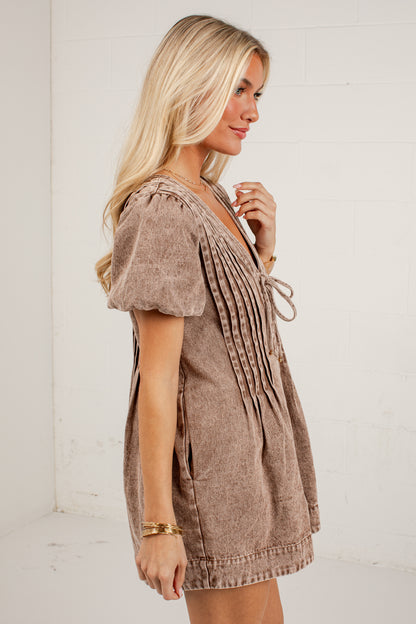 A woman wearing a brown short-sleeve mini dress with pleated detailing and tie-front laces, standing against a white brick wall.