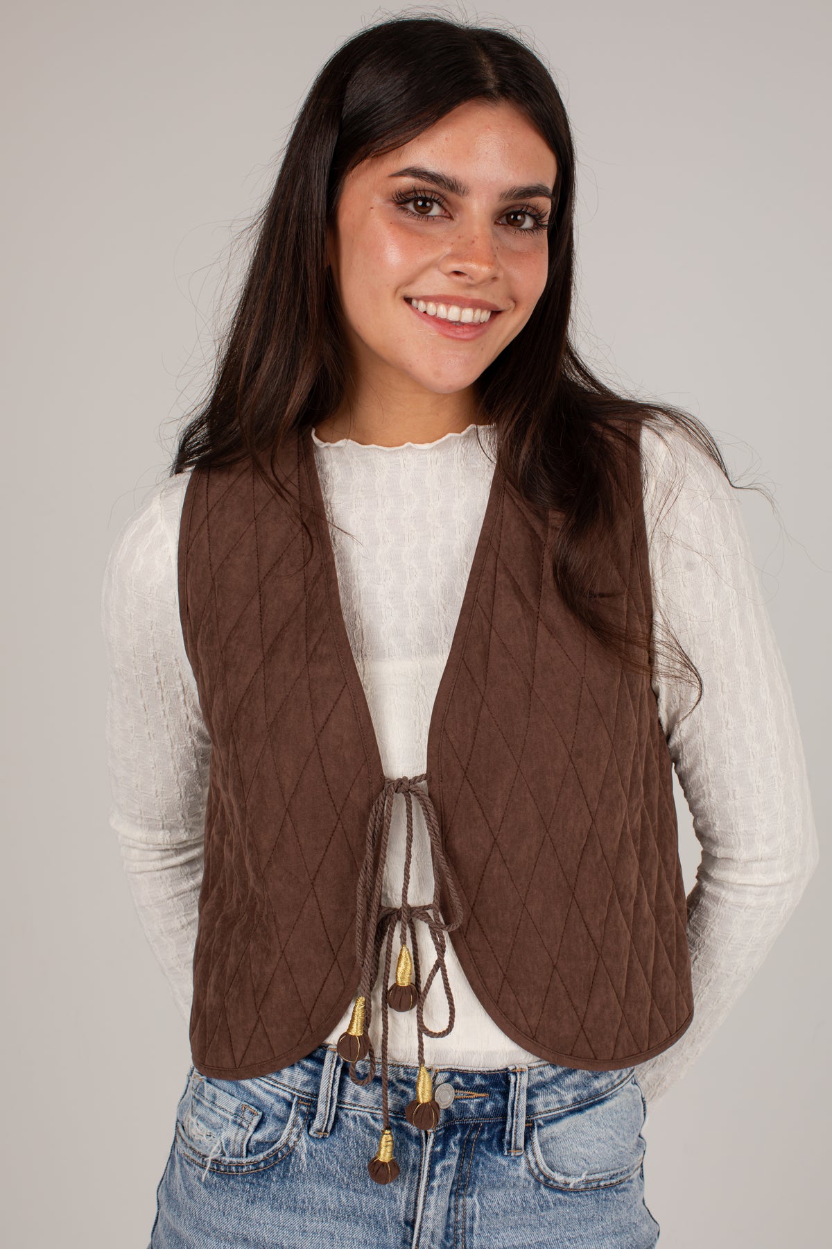 A woman wearing a brown diamond-quilted vest over a white long-sleeve top and blue jeans, photographed against a plain light gray studio background.