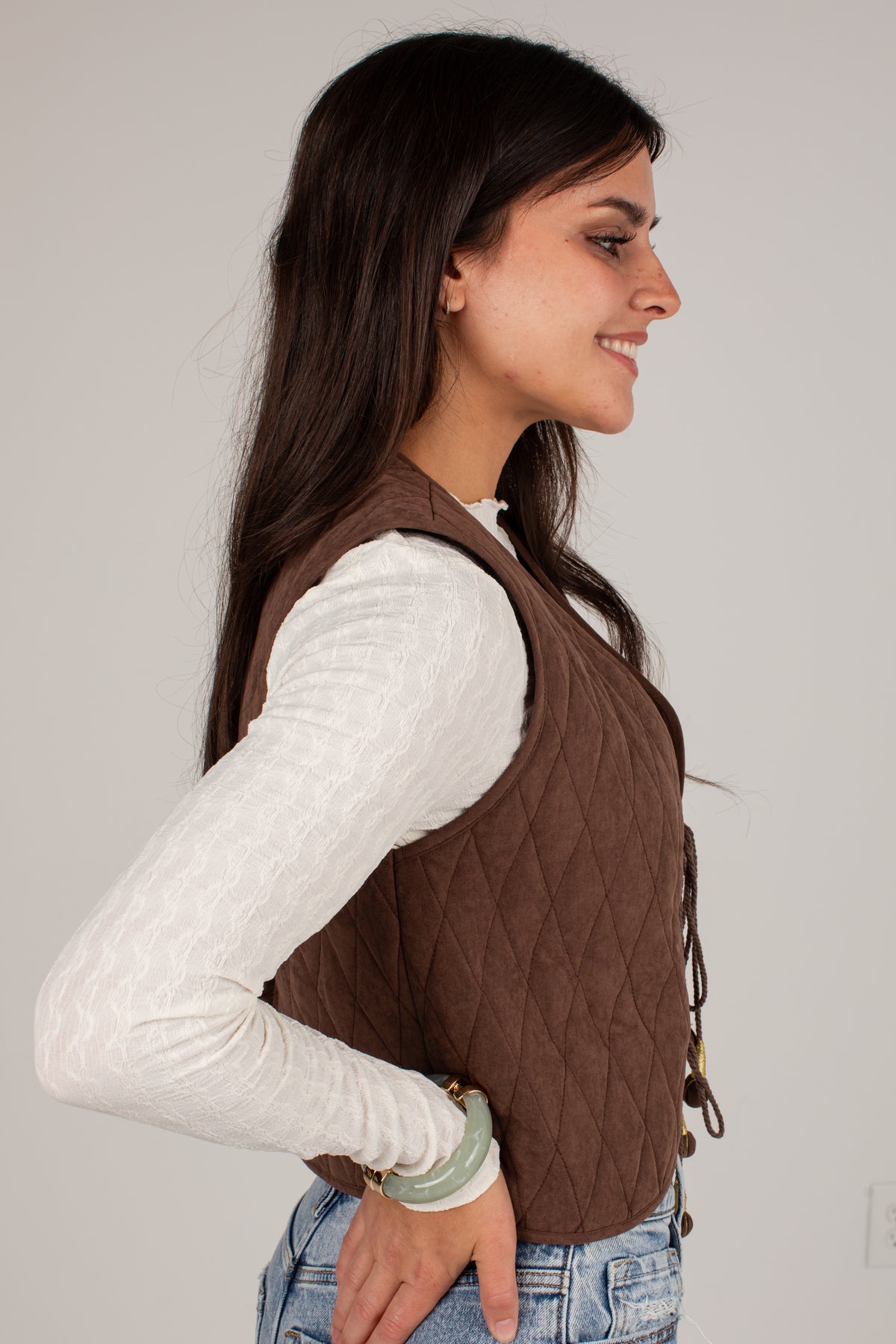 A woman shown in profile wearing a brown diamond-quilted sleeveless vest over a white long-sleeve top and blue jeans, against a plain light gray studio background.