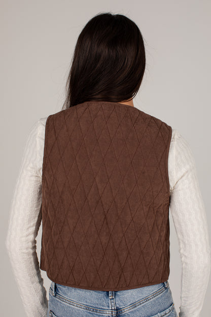 A person shown from the back wearing a brown diamond-quilted sleeveless vest over a white ribbed long-sleeve top and light blue jeans, against a plain light gray studio background.