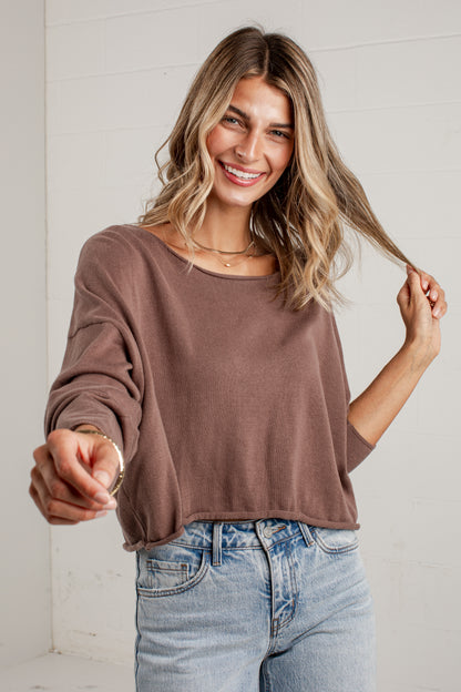 A woman wearing a brown long-sleeve cropped knit top and light blue jeans, smiling and pointing toward the camera against a white paneled wall.
