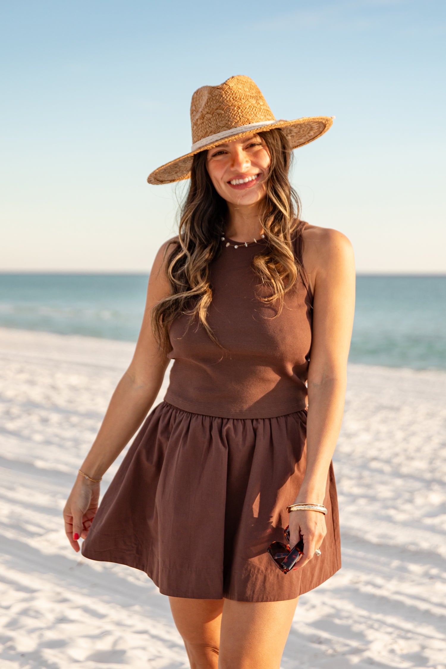 Woman in a brown dress and hat standing on a sandy beach with ocean view