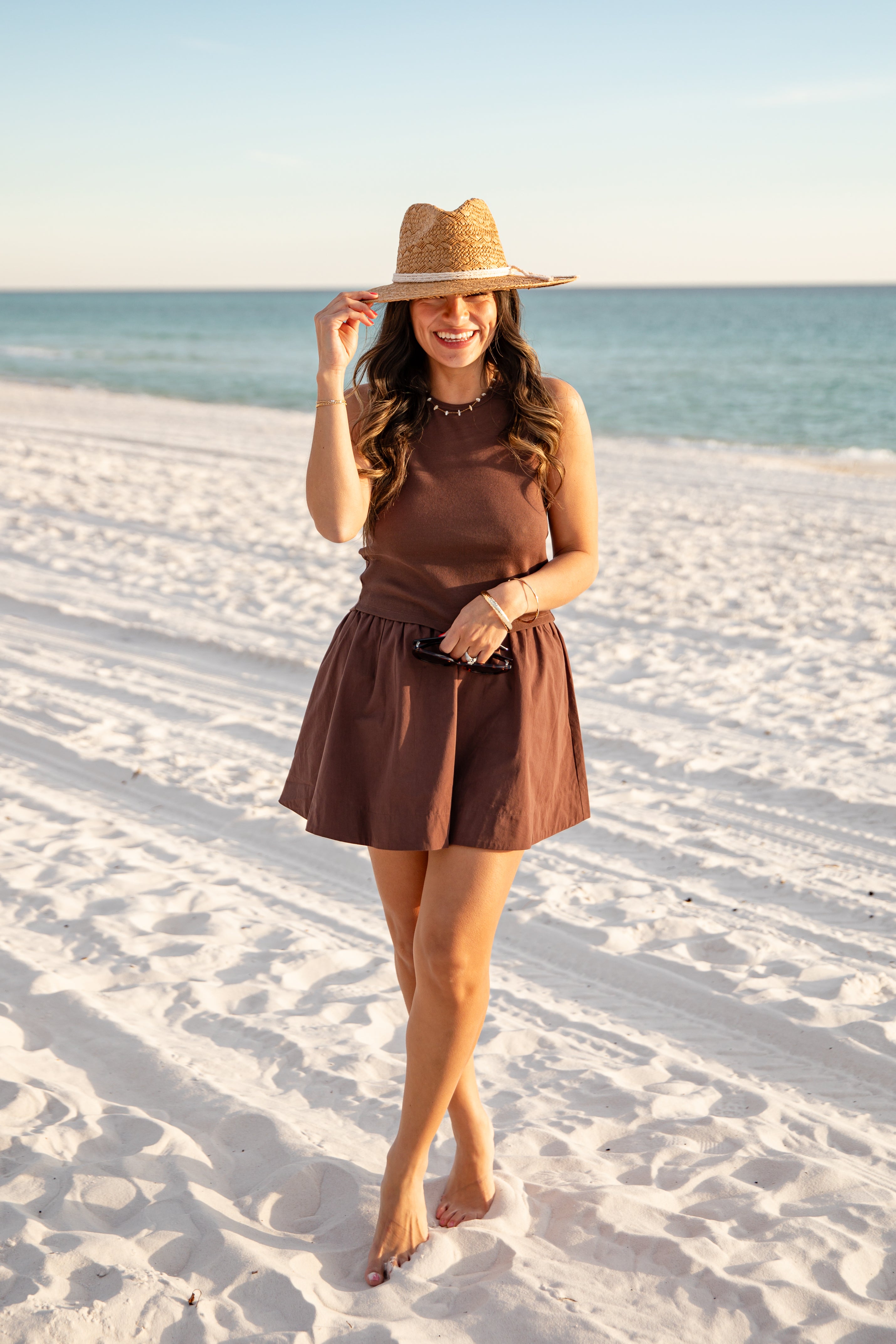 Woman in a brown dress and hat standing on a sandy beach with ocean in the background