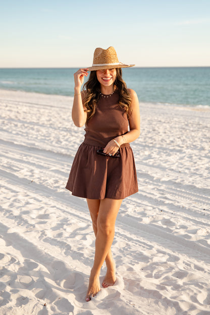 Woman in a brown dress and hat standing on a sandy beach with ocean in the background