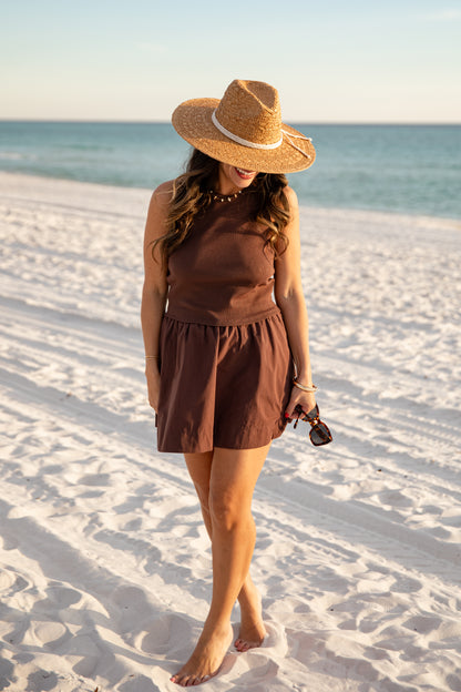 Woman in a brown dress and hat standing on a sandy beach with ocean in the background