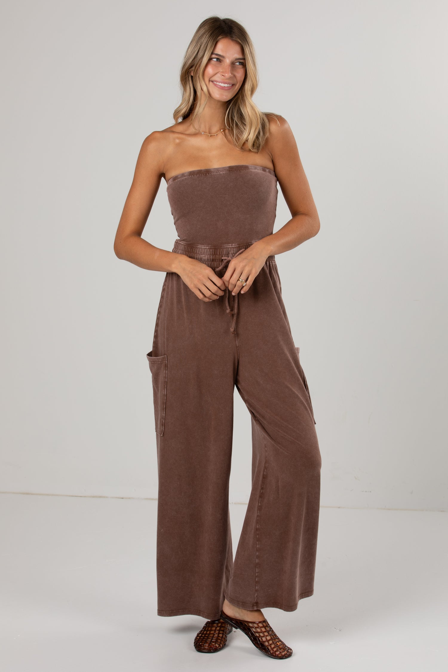 A woman modeling a brown strapless wide-leg jumpsuit with a matching tie belt at the waist, photographed against a plain light gray studio background.